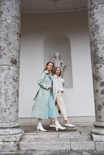 Two women in stylish pastel coats and dresses pose near stone pillars, complemented by white ankle boots, in an outdoor setting with a statue.
