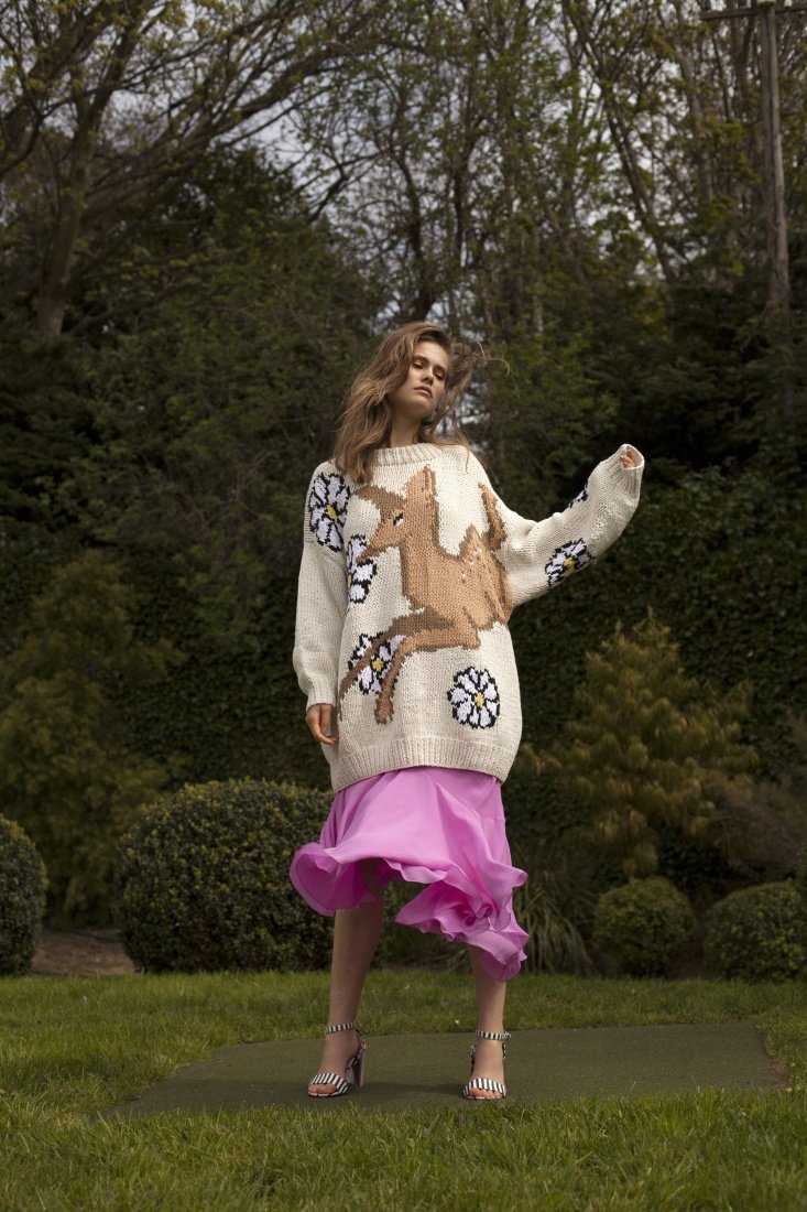 A woman stands in a garden wearing a cream sweater with a large deer design, paired with a flowing pink skirt and striped heels.