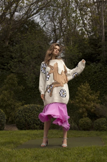 A woman stands in a garden wearing a cream sweater with a large deer design, paired with a flowing pink skirt and striped heels.