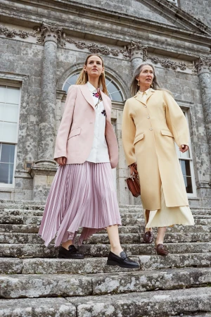 Two women in stylish coats and loafers walk down stone steps outside a grand building, showcasing fashion in a historic setting.