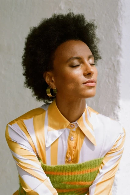 Portrait of a woman with dark curly hair, wearing a striped yellow and white shirt with gold hoop earrings, eyes closed in sunlight.