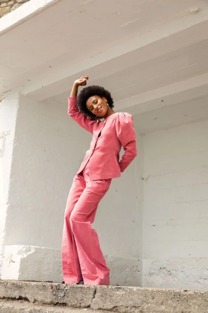 Portrait of a smiling woman with natural hair in a pink tailored suit standing outdoors on a concrete structure in sunlight.
