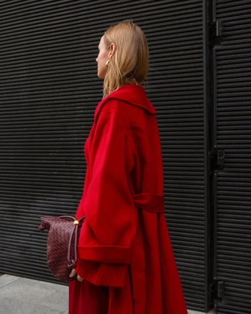 Woman wearing a bright red belted wool coat and holding a dark red leather woven handbag against a dark ribbed metal wall.