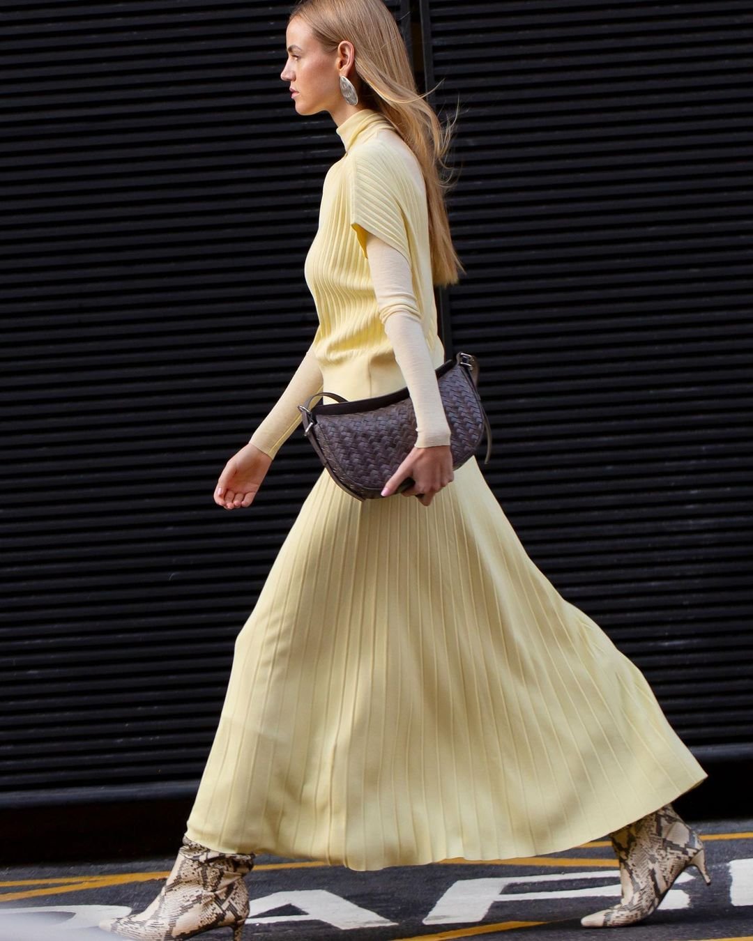 Fashionable woman in a yellow dress and snake print boots walks confidently against a dark corrugated background, showcasing street style.