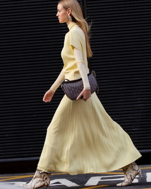 Fashionable woman in a yellow dress and snake print boots walks confidently against a dark corrugated background, showcasing street style.