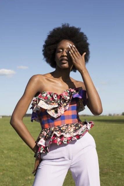 A woman with an afro, wearing a colorful floral and plaid ruffled top and light purple pants, stands outdoors in a grassy field.