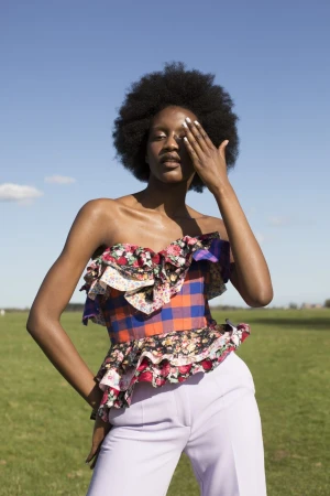 A woman with an afro, wearing a colorful floral and plaid ruffled top and light purple pants, stands outdoors in a grassy field.