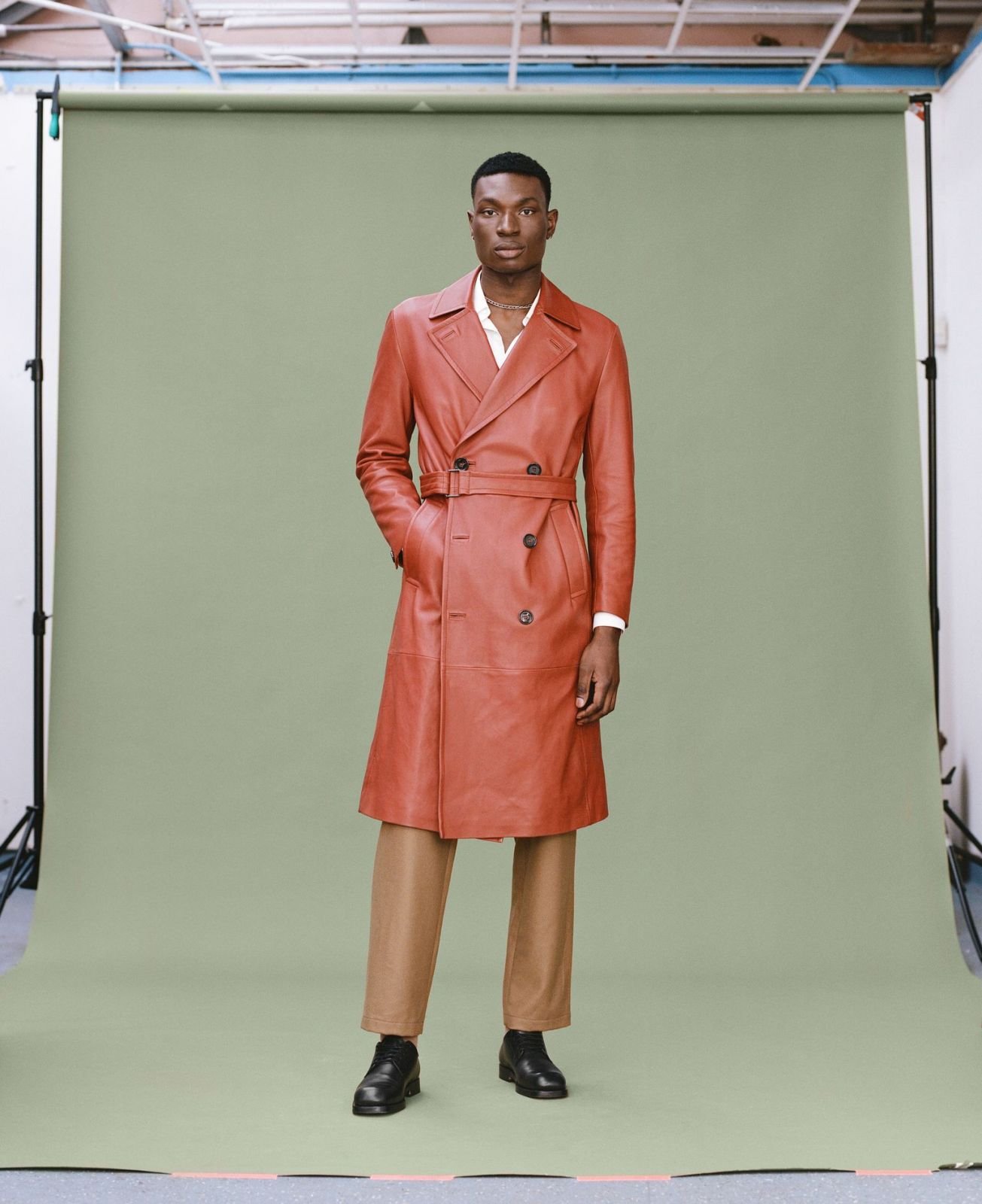 Full-length shot of an adult male wearing a long, double-breasted, brown leather coat with belt, tan pants, and black leather shoes.