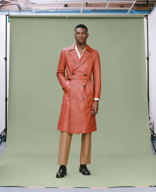 Full-length shot of an adult male wearing a long, double-breasted, brown leather coat with belt, tan pants, and black leather shoes.