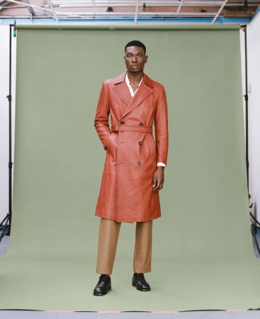 Full-length shot of an adult male wearing a long, double-breasted, brown leather coat with belt, tan pants, and black leather shoes.