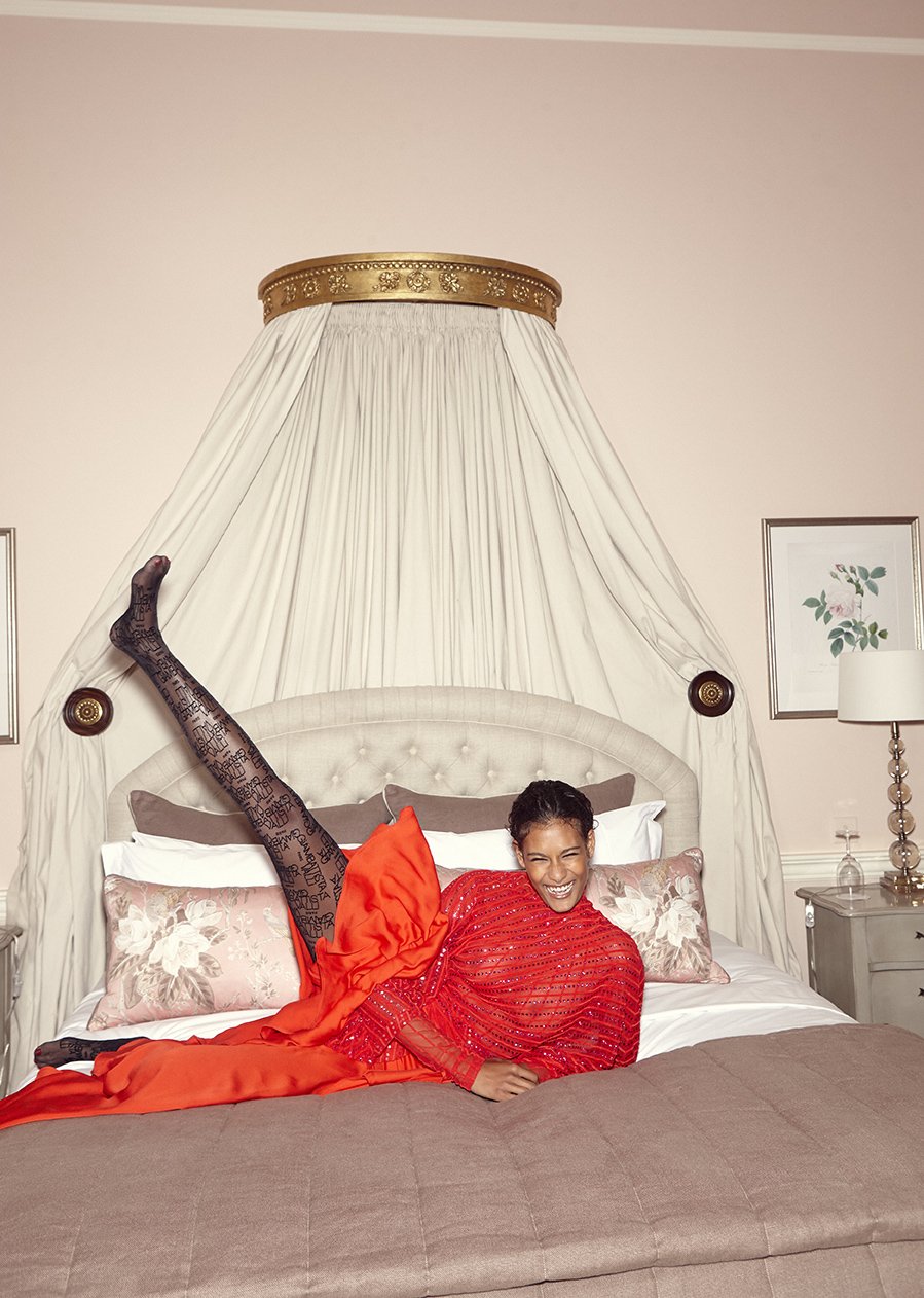 Woman in a red dress and patterned stockings laying on a bed with her legs up in the air in an indoor bedroom setting.