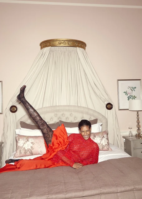 Woman in a red dress and patterned stockings laying on a bed with her legs up in the air in an indoor bedroom setting.