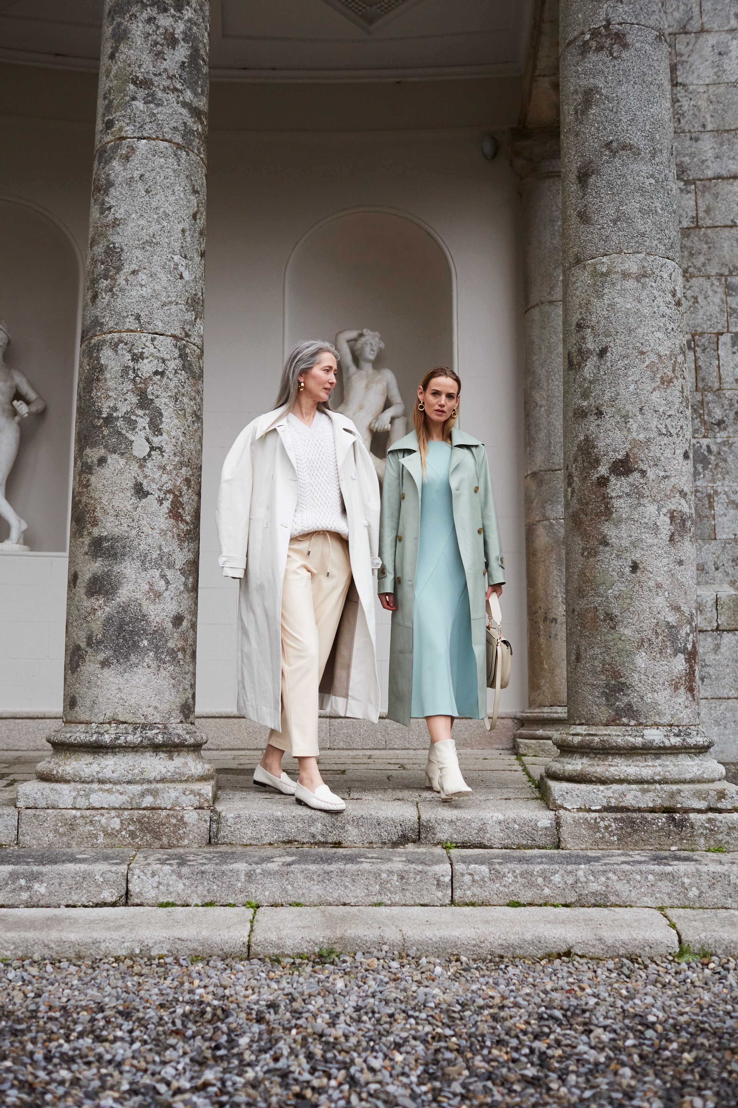 Two stylish women in coats walk on stone steps near a classical building with statues, dressed in fashionable neutral colors.