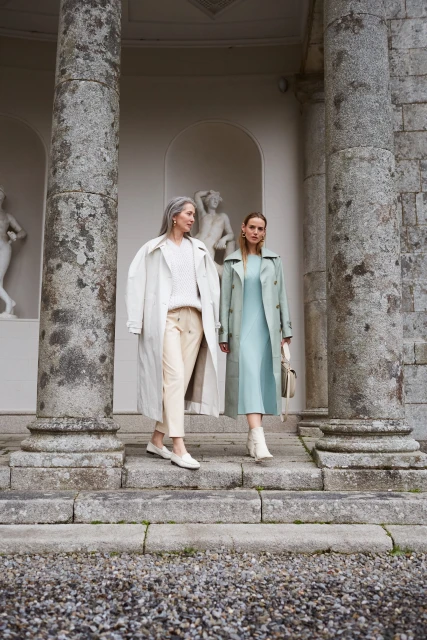 Two stylish women in coats walk on stone steps near a classical building with statues, dressed in fashionable neutral colors.