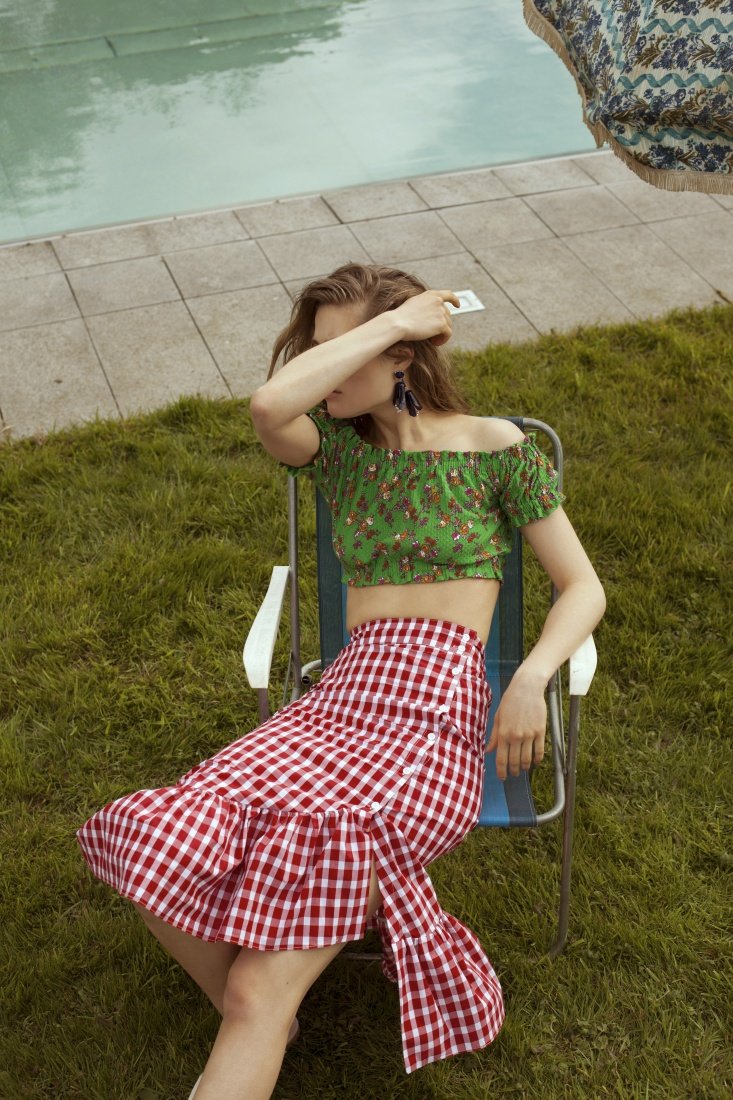 Woman in a green crop top and red gingham skirt sits on a blue and white chair on green grass near a tiled pool on a sunny day.