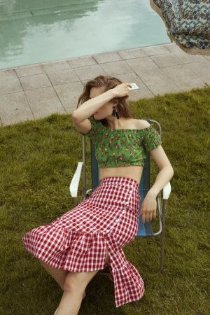 Woman in a green crop top and red gingham skirt sits on a blue and white chair on green grass near a tiled pool on a sunny day.