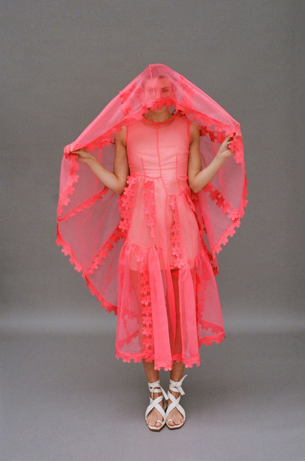 A female model showcases a pink floral dress with a matching veil and white tie-up sandals against a neutral grey backdrop in a studio setting.