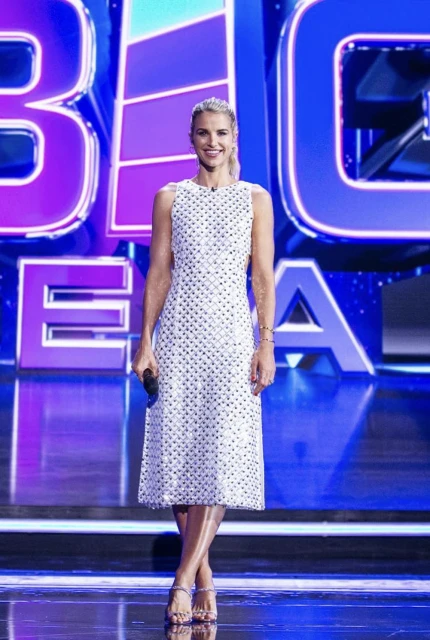 Adult woman in white dress standing on stage with large blue 'BIG IDEA' text, holding microphone, full body shot, bright stage lighting.