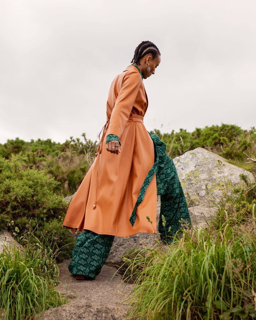A woman in a long, tan jacket and patterned pants walks uphill on a stone path surrounded by green plants under an overcast sky.