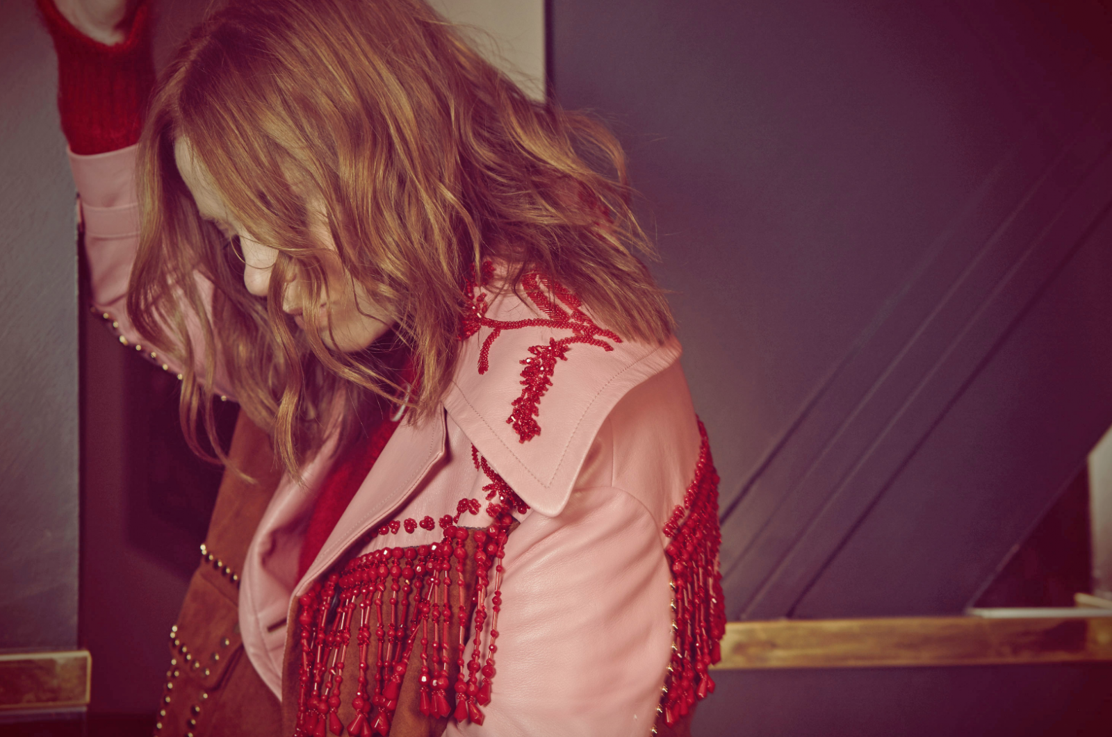 Woman in a pink jacket with red beaded fringe, facing away from the viewer with long hair. Shoulder and back detail, indoors.