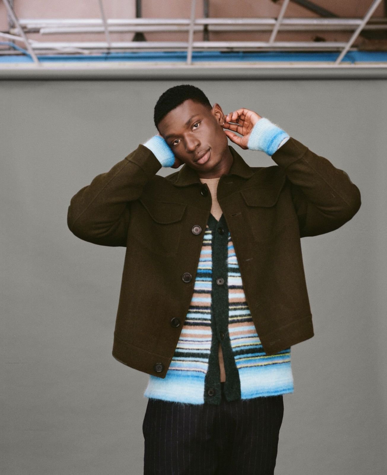 Studio shot of a Black male model in a green jacket and colorful striped cardigan, gazing at the camera with neutral expression.
