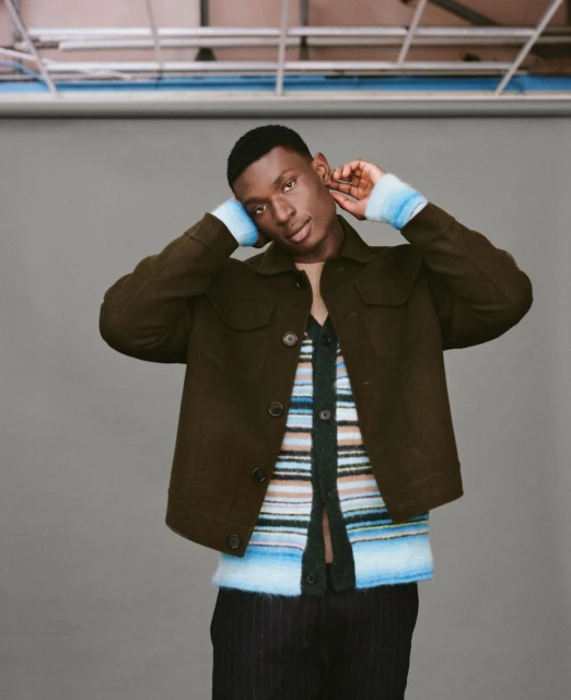 Studio shot of a Black male model in a green jacket and colorful striped cardigan, gazing at the camera with neutral expression.