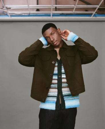 Studio shot of a Black male model in a green jacket and colorful striped cardigan, gazing at the camera with neutral expression.