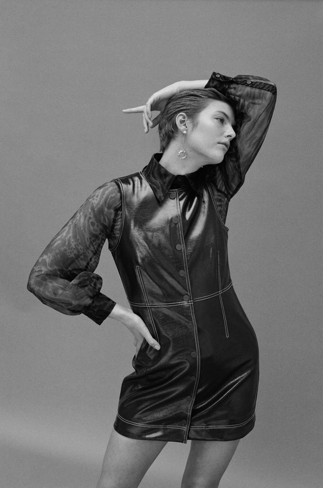 Black and white studio shot of a female model in a black faux leather dress and sheer black top, striking a fashion pose.
