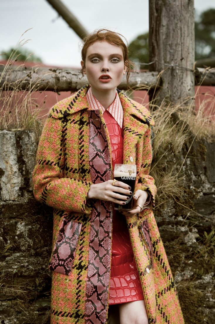 Fashion editorial shot of a red-haired female model in a plaid coat holding a pint of Guinness in front of a stone wall and field.