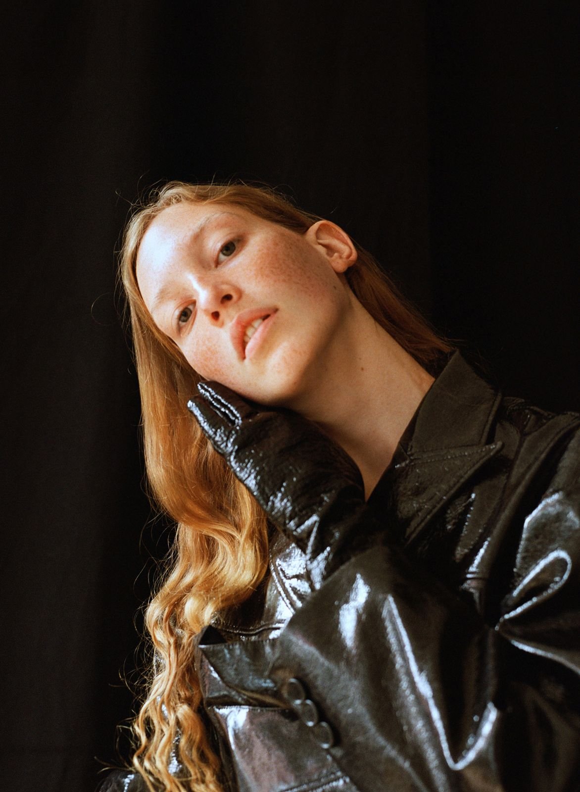Headshot of a fair-skinned adult female with long wavy red hair wearing a shiny black jacket and gloves against a black background.
