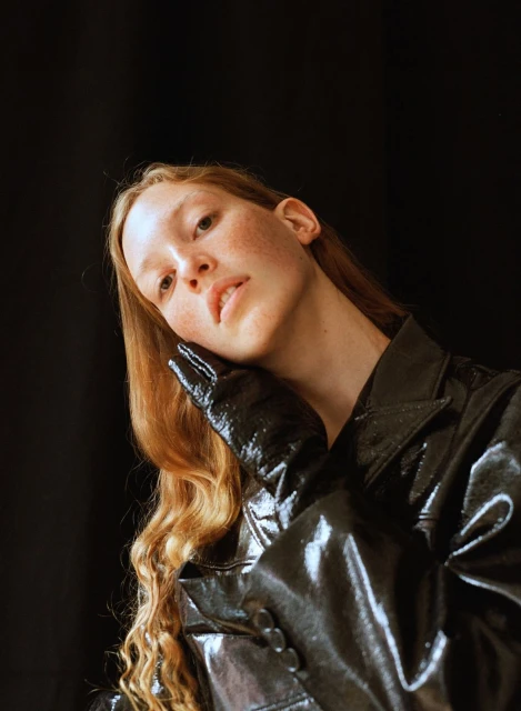 Headshot of a fair-skinned adult female with long wavy red hair wearing a shiny black jacket and gloves against a black background.