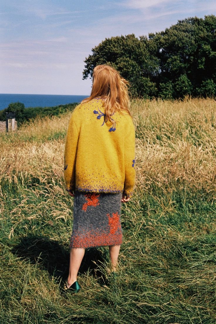 Back view of a woman with long red hair in a yellow patterned sweater and grey skirt standing in a field of tall grass.