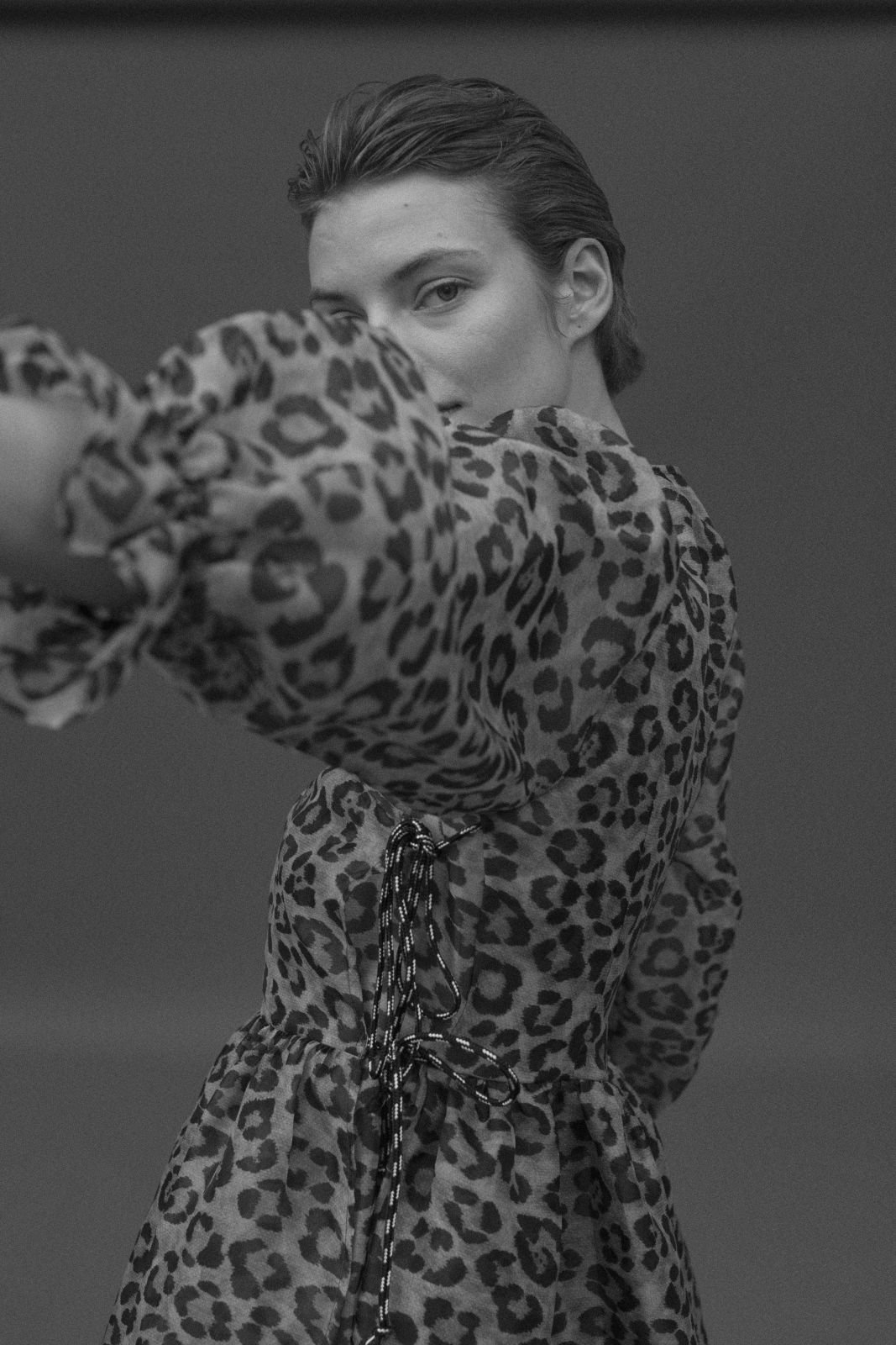 Monochrome portrait of a stylish young adult woman in an animal-print dress, posing in front of a gray backdrop with soft studio lighting.