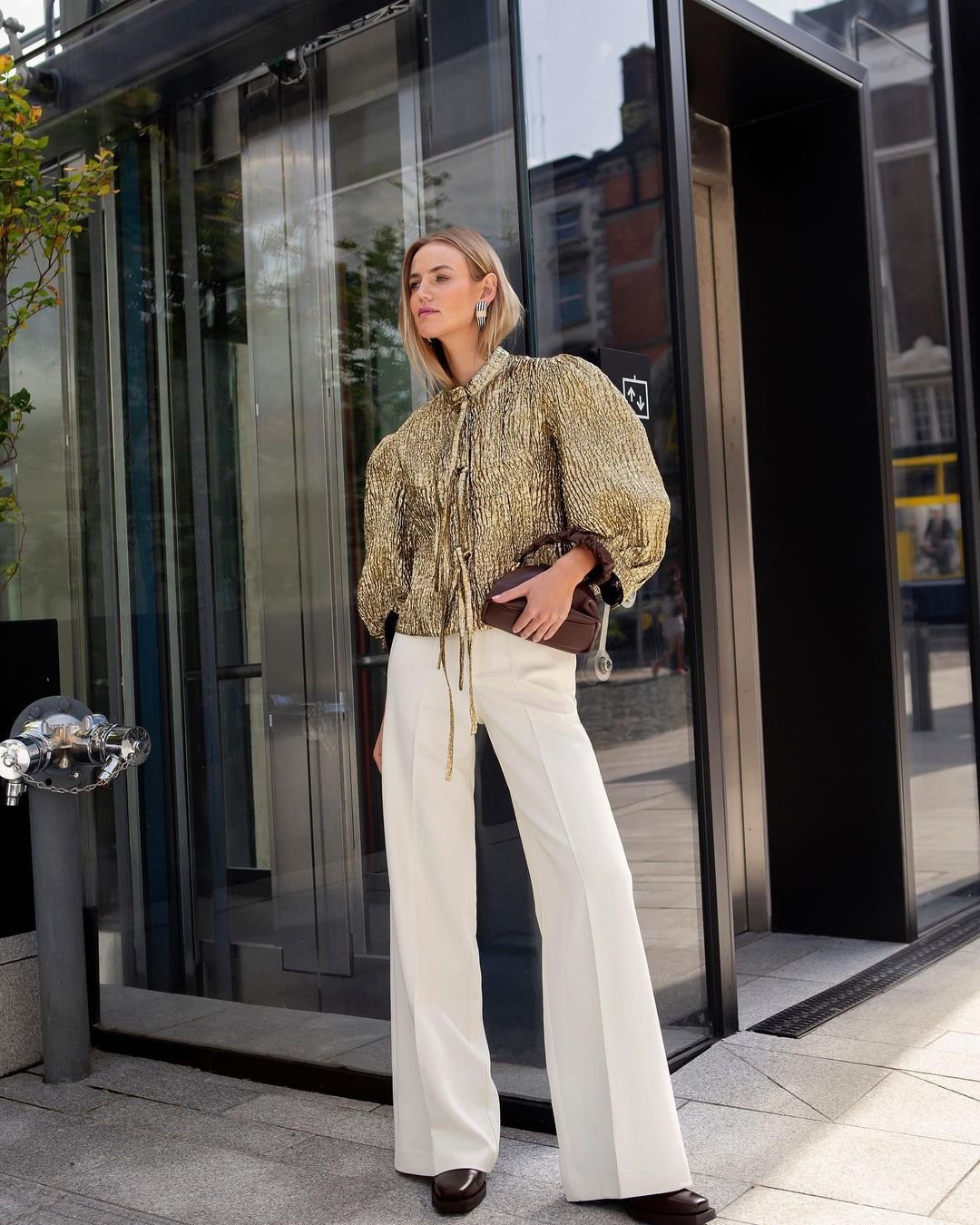 Blonde woman in gold lamé blouse with oversized sleeves and white wide-leg trousers, posing outdoors with a small brown handbag.
