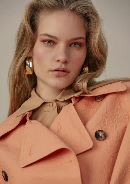 Close-up studio shot of a young adult woman with wavy blonde hair wearing gold hoop earrings and a pink trench coat.
