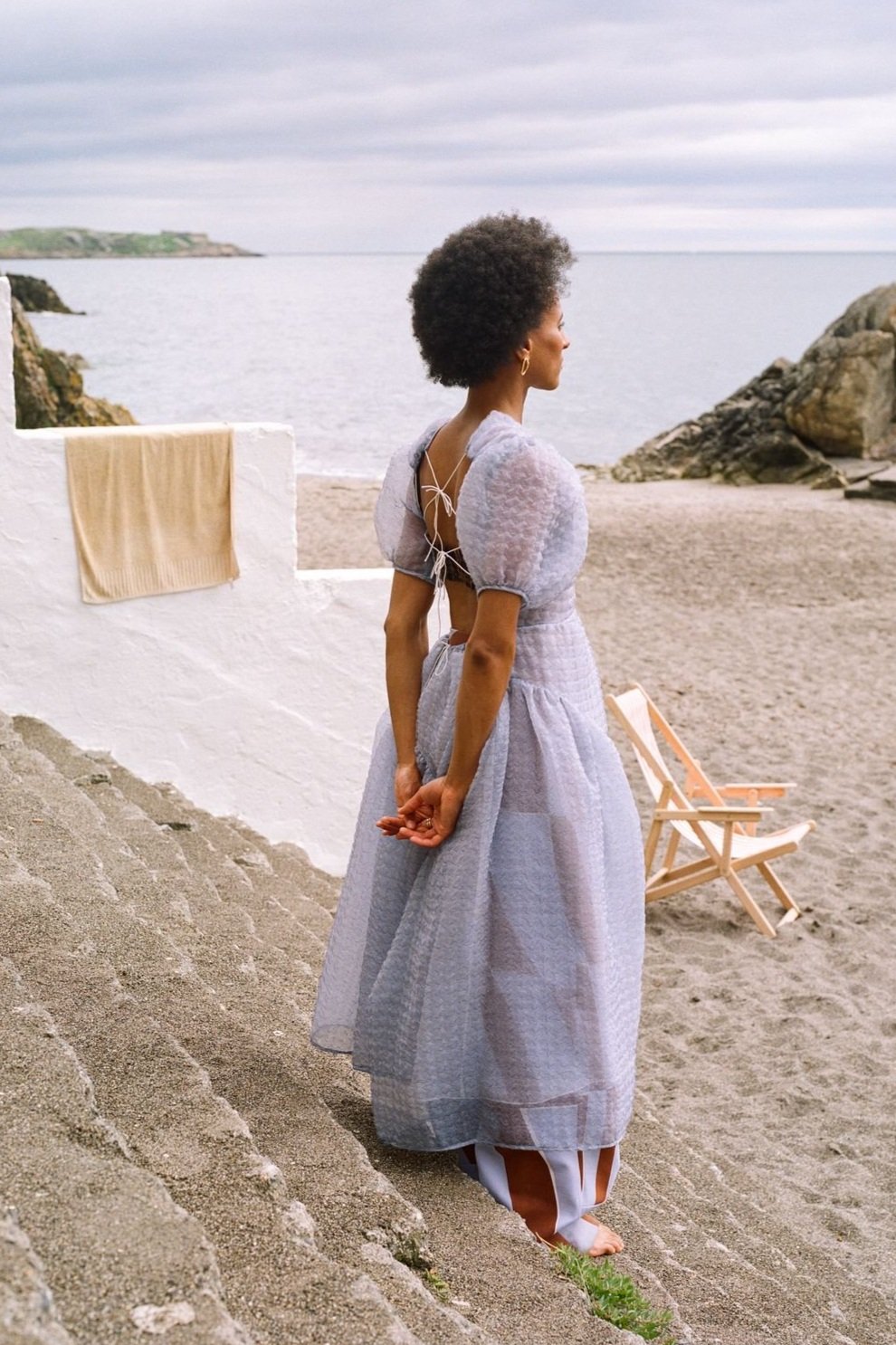 A lone woman in a flowing dress stands on stone steps at a beachside; the light airy fabric complements the dreamy seaside ambiance.
