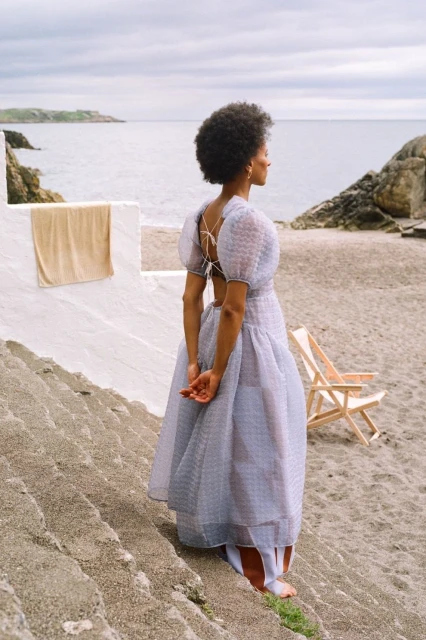 A lone woman in a flowing dress stands on stone steps at a beachside; the light airy fabric complements the dreamy seaside ambiance.