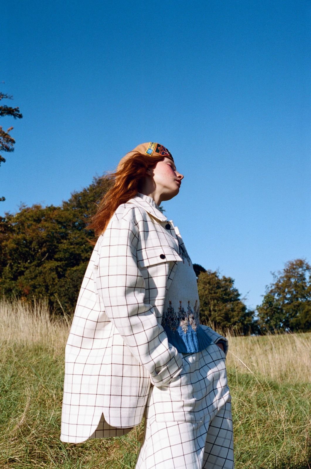 An outdoor shot of an adult woman in a patterned coat and pants, looking up with trees and blue sky in the background on a sunny day.