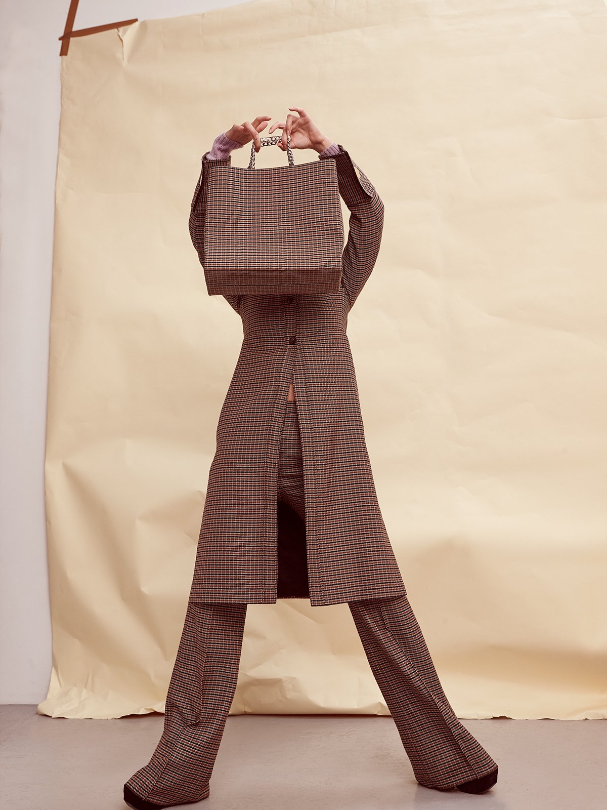 Fashionable woman in brown plaid suit poses with a plaid shopping bag in front of her face in a brightly lit photo studio setting.