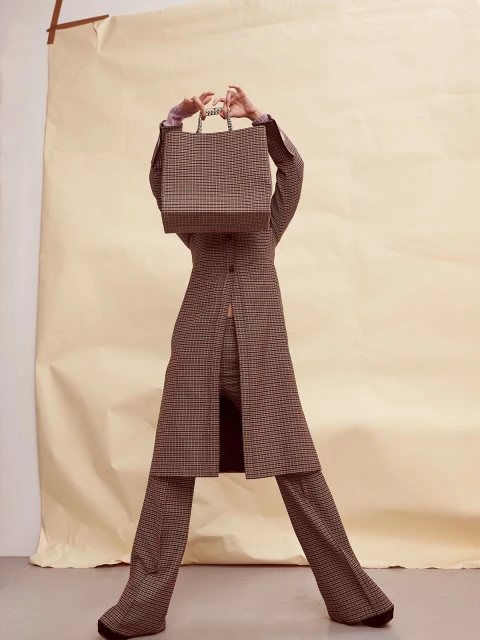 Fashionable woman in brown plaid suit poses with a plaid shopping bag in front of her face in a brightly lit photo studio setting.