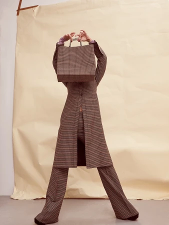 Fashionable woman in brown plaid suit poses with a plaid shopping bag in front of her face in a brightly lit photo studio setting.