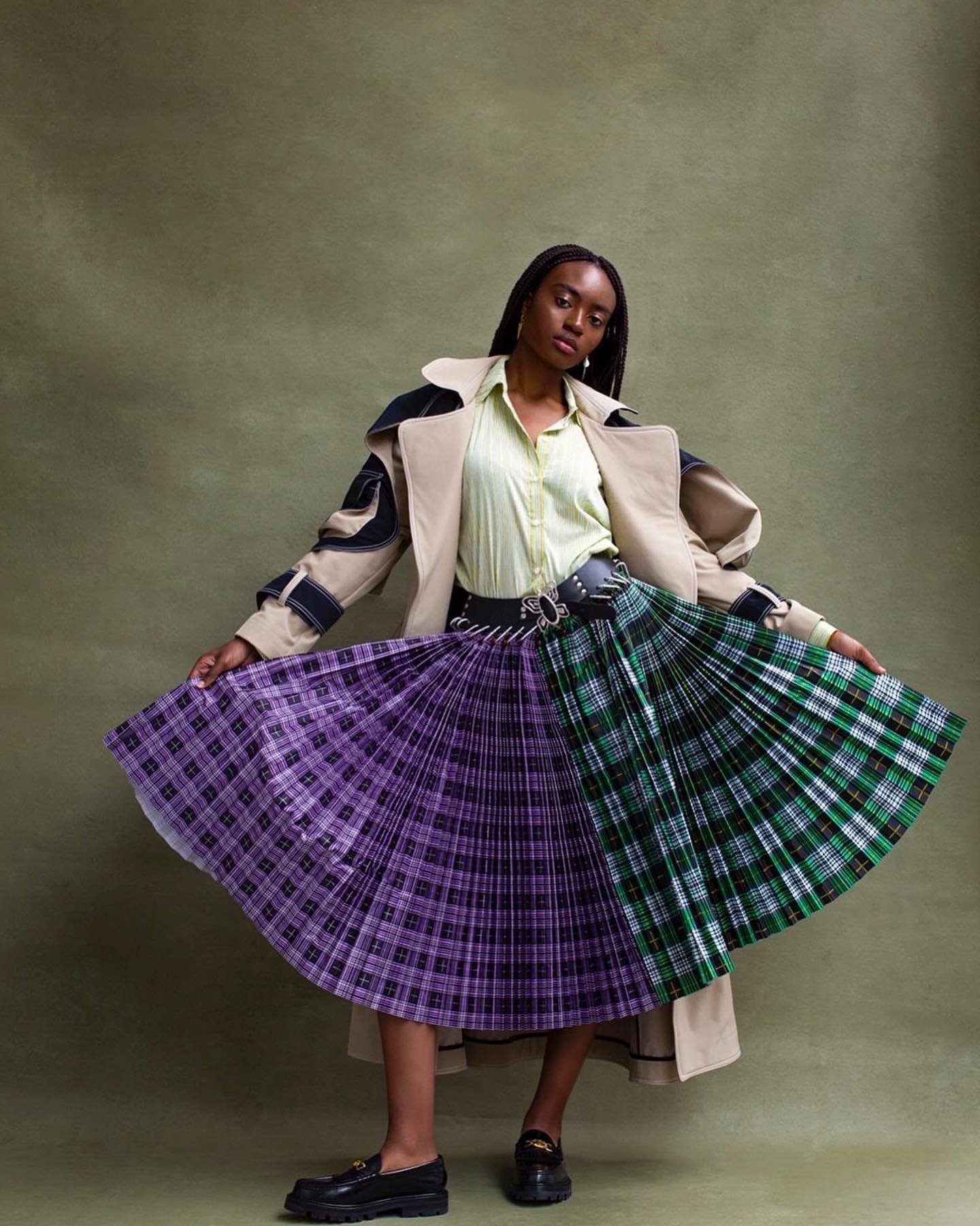 An African-American woman poses in a studio wearing a beige trench coat, a light-green shirt, and a vibrant plaid skirt.