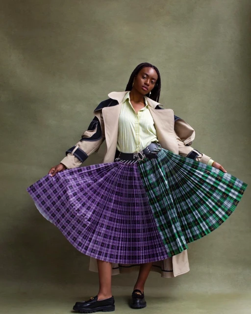 An African-American woman poses in a studio wearing a beige trench coat, a light-green shirt, and a vibrant plaid skirt.