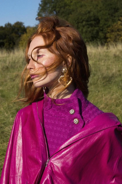 Red-haired woman in magenta coat and oversized earrings posing in field with green grass and trees under a blue sky on a sunny day.