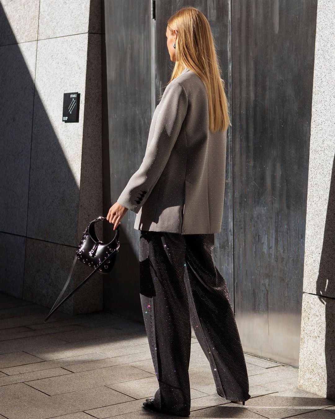 Adult woman in a grey blazer and sparkling black wide-leg pants walks on a sidewalk, holding a small black studded purse.