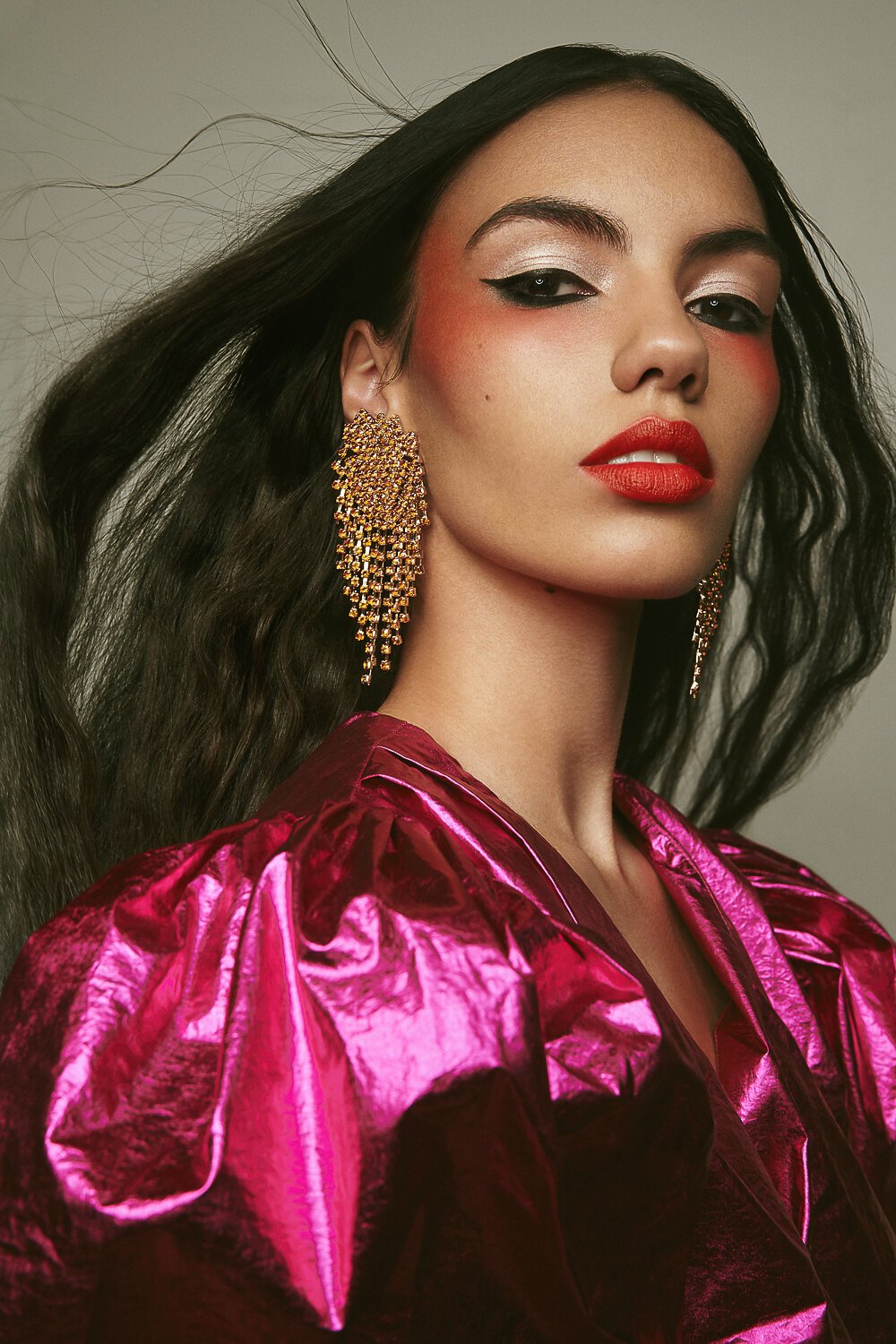 Close-up portrait of a female model with long dark hair, vibrant red lipstick, and large gold earrings, wearing a shiny pink blouse.