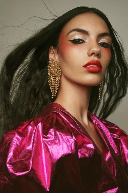 Close-up portrait of a female model with long dark hair, vibrant red lipstick, and large gold earrings, wearing a shiny pink blouse.