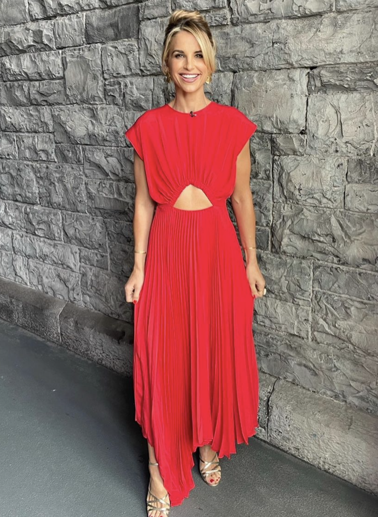 A woman with blonde hair smiles, wearing a red dress with gold sandals, standing against a gray brick wall, photographed in portrait.