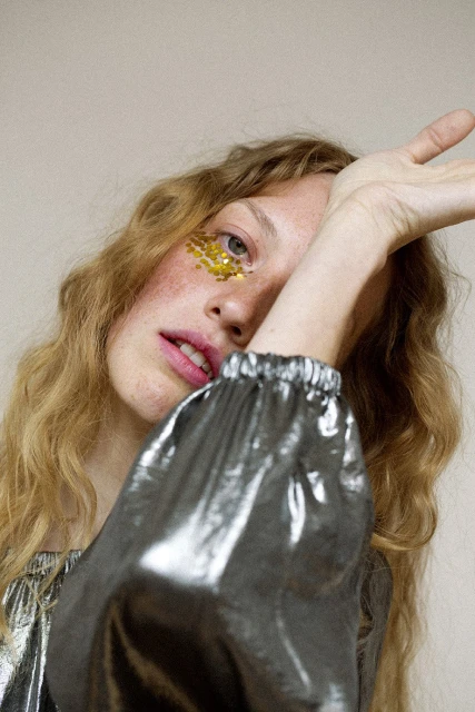 Portrait of a stylish young adult woman with gold glitter makeup on her face, wearing a silver shiny top, posing indoors.