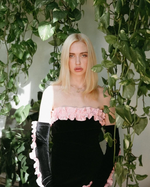 A blonde woman with long velvet gloves poses in front of a green leafy wall. She's wearing a black dress with pink ruffles.